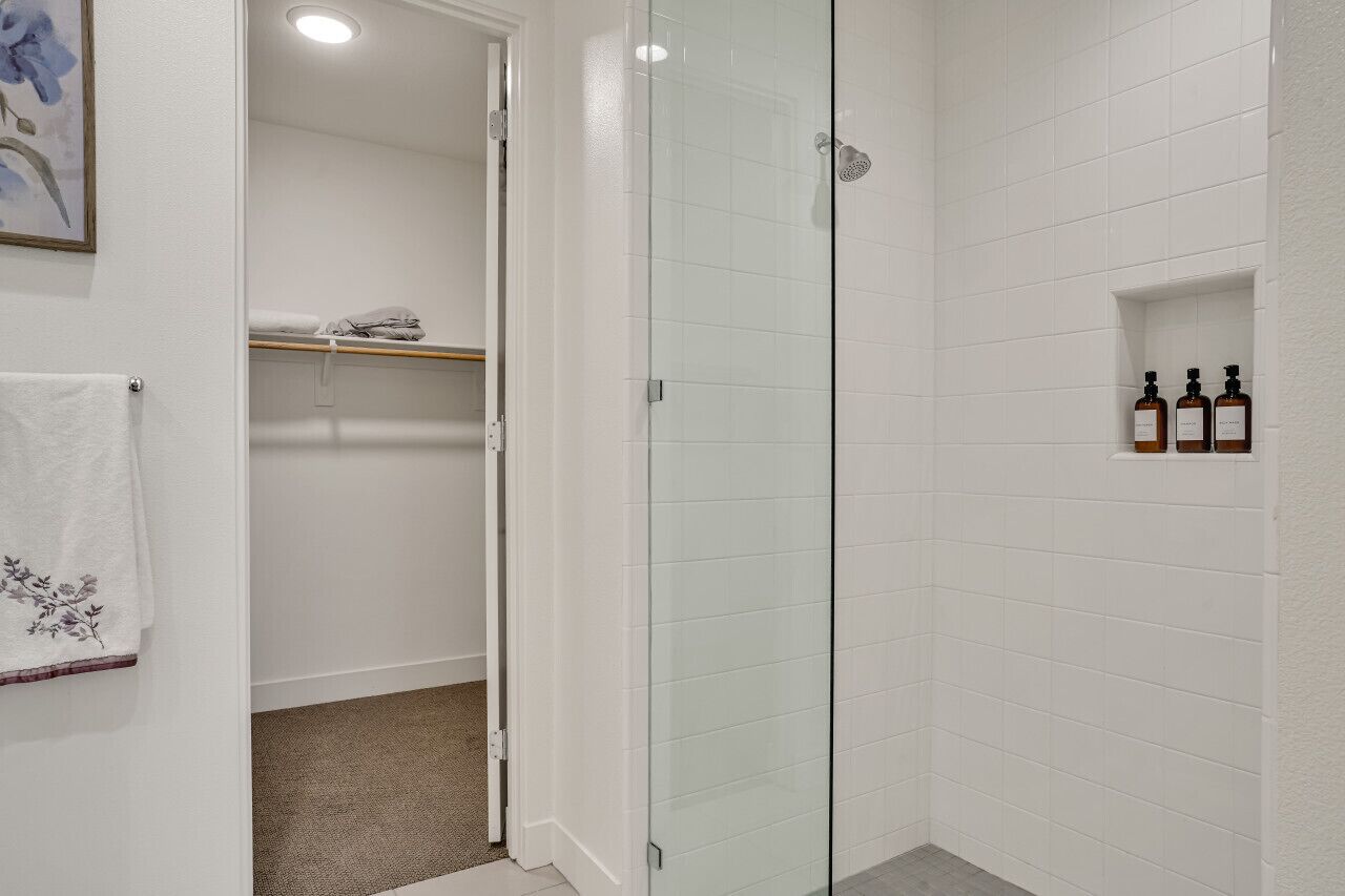 A bathroom with white tiled walls, a glass shower enclosure, a towel rack with a white towel, and a shelf with two bottles.
