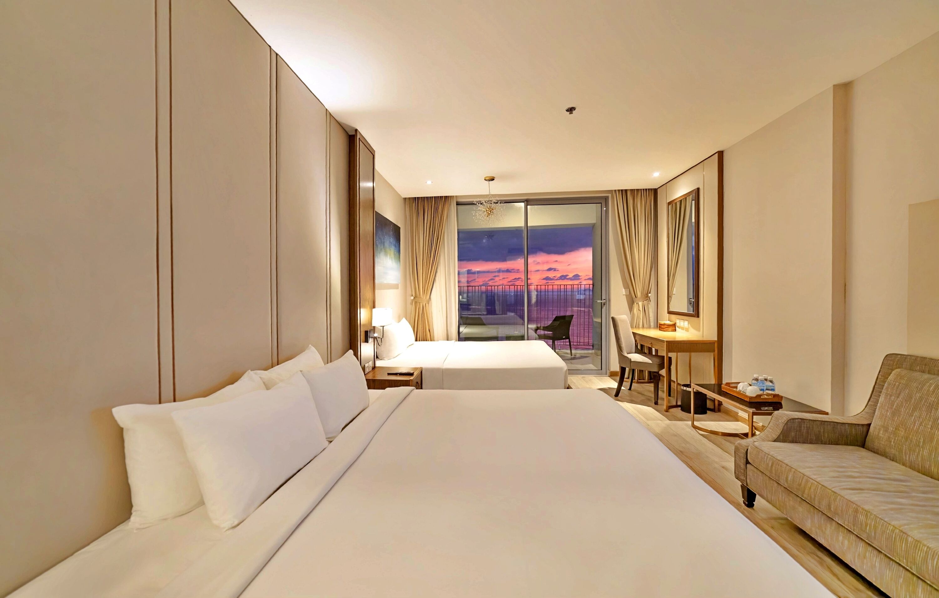 A hotel room with a large bed, a sofa, a desk with a chair, and a view of a sunset through a glass door.