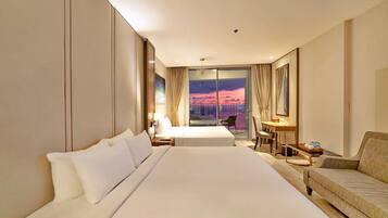 A hotel room with a large bed, a sofa, a desk with a chair, and a view of a sunset through a glass door.