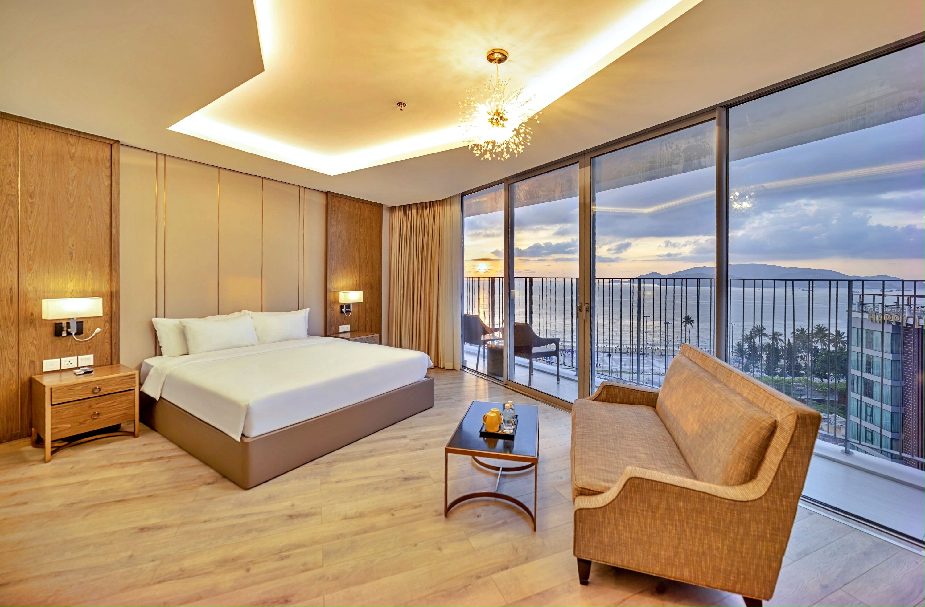A modern hotel room with a large bed, a sofa, a small table, and a balcony with a city view.