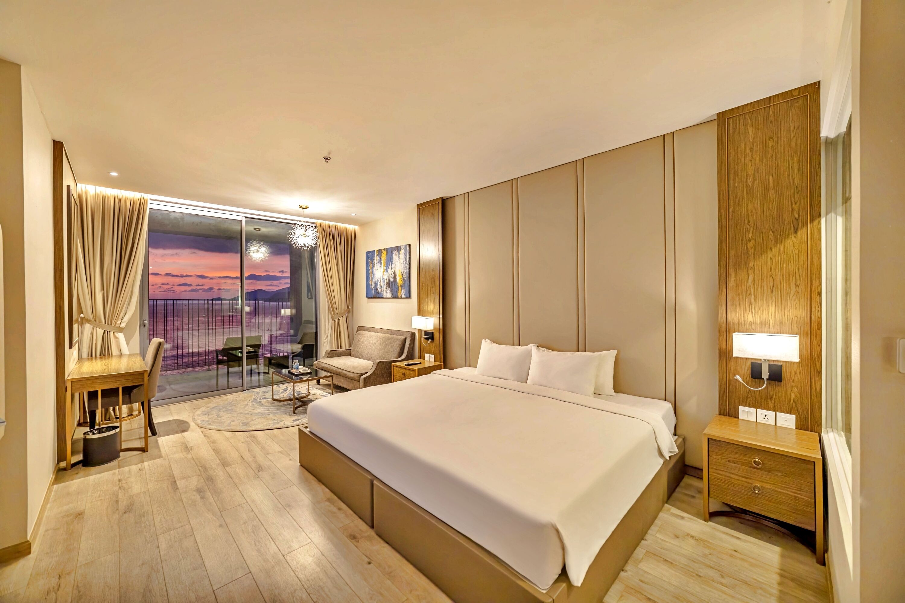 A hotel room with a large bed, a desk, a chair, a small table, a nightstand, and a view of the sunset through a glass door.