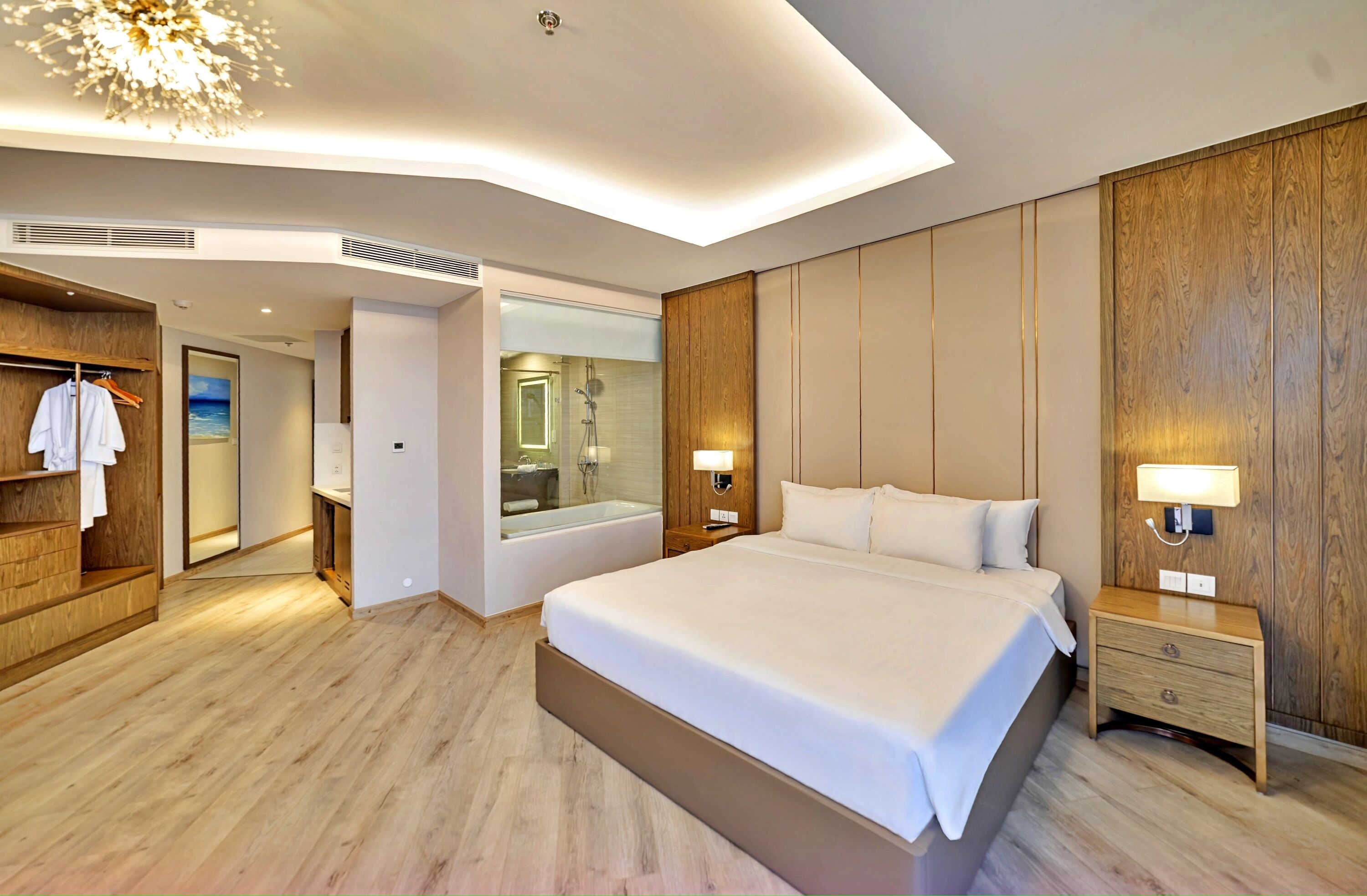 A modern hotel room with a large bed, wooden paneling, a bathroom with a bathtub, and a view of the sea through a glass door.