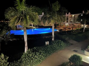 A nighttime view of a hotel with a swimming pool, palm trees, and a well-lit outdoor area.