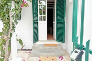 Un edificio blanco con contraventanas verdes, un escalón de hormigón que lleva a la entrada y un felpudo de madera pequeño.