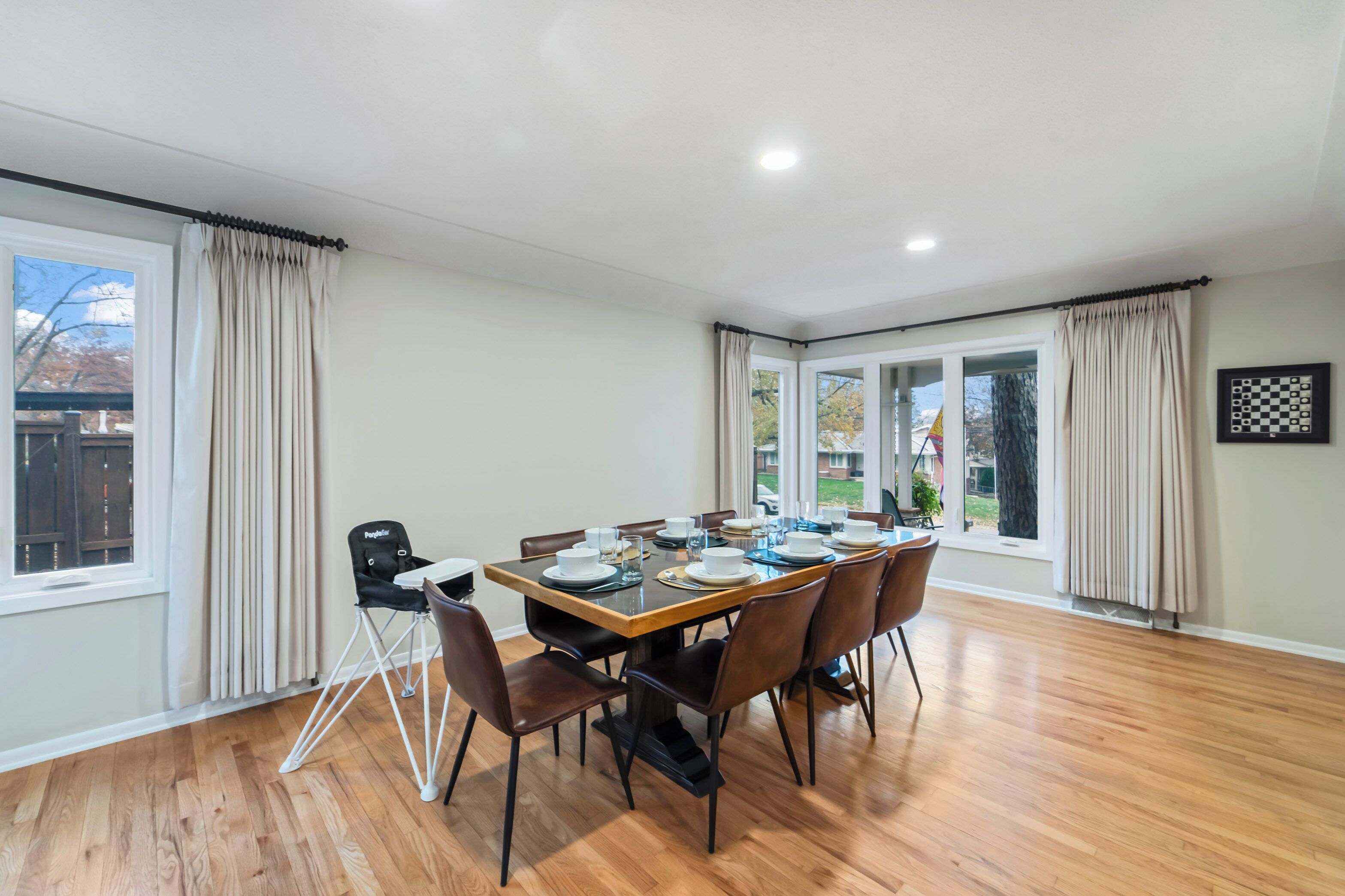 Un coin repas avec une table en bois dressée pour six personnes, entourée de chaises, et offrant une vue sur un jardin grâce à de grandes fenêtres.