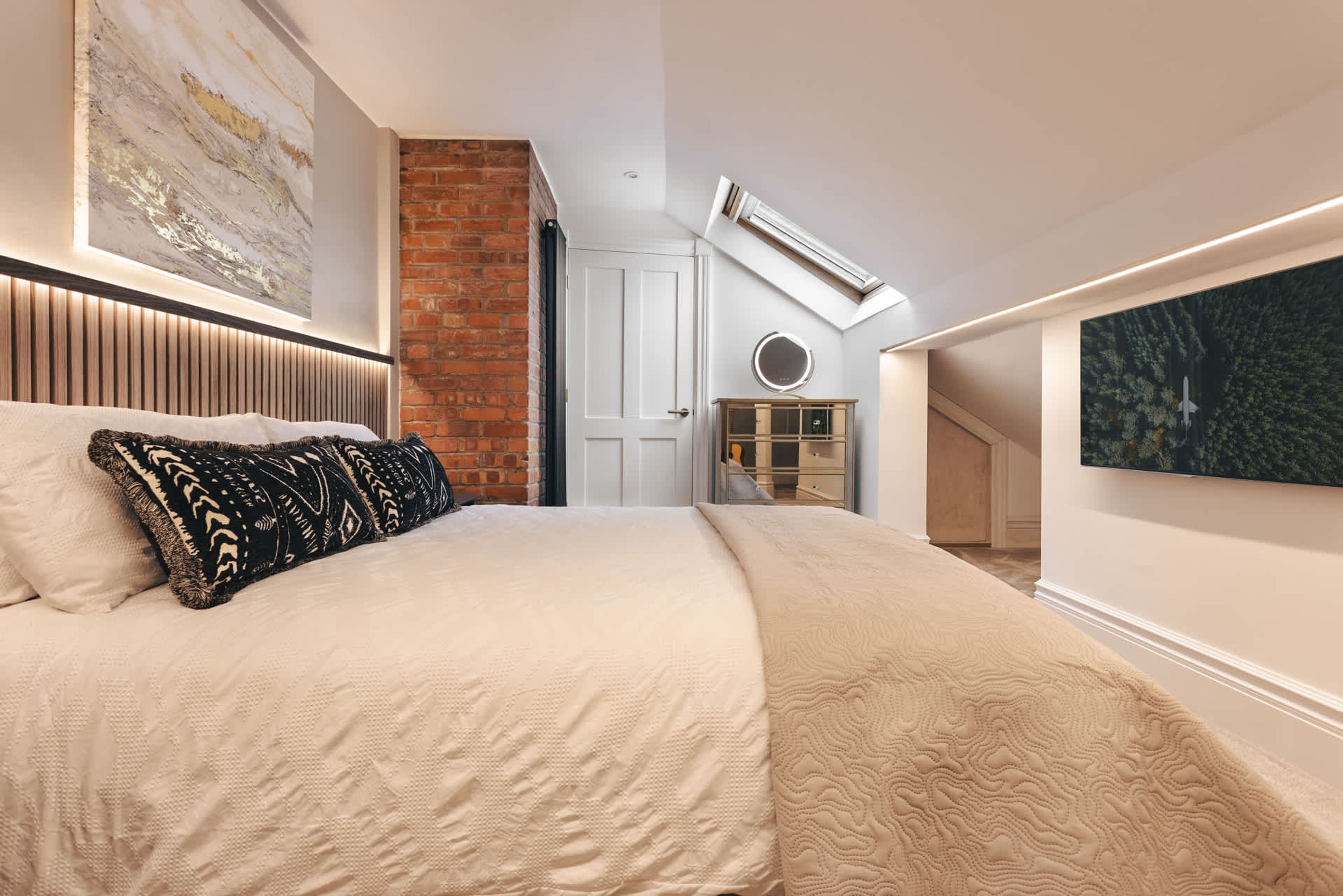 A bedroom with a large bed, a brick wall, a skylight, and a door leading to another room.