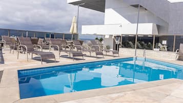 Uma piscina no terraço com espreguiçadeiras, vista desobstruída da cidade e um edifício moderno com paredes revestidas de azulejos.