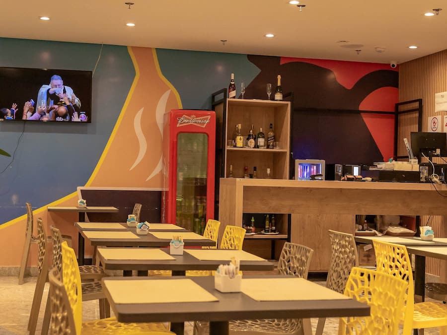 A restaurant with tables and chairs, a television screen, and a shelf with bottles.