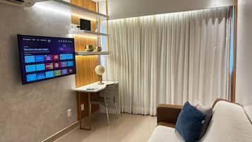 Uma sala de estar moderna com uma TV de tela plana na parede exibindo diversos aplicativos, uma escrivaninha com abajur globo, um sofá marrom com travesseiro azul e uma grande janela com cortinas brancas.