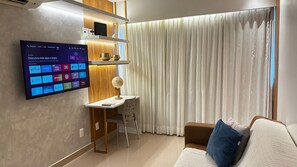 A modern living room with a flat-screen TV mounted on the wall displaying various apps, a desk with a globe lamp, a brown sofa with a blue pillow, and a large window with white curtains. - Flats no Beach Class Residence por Yolo  (Recife)