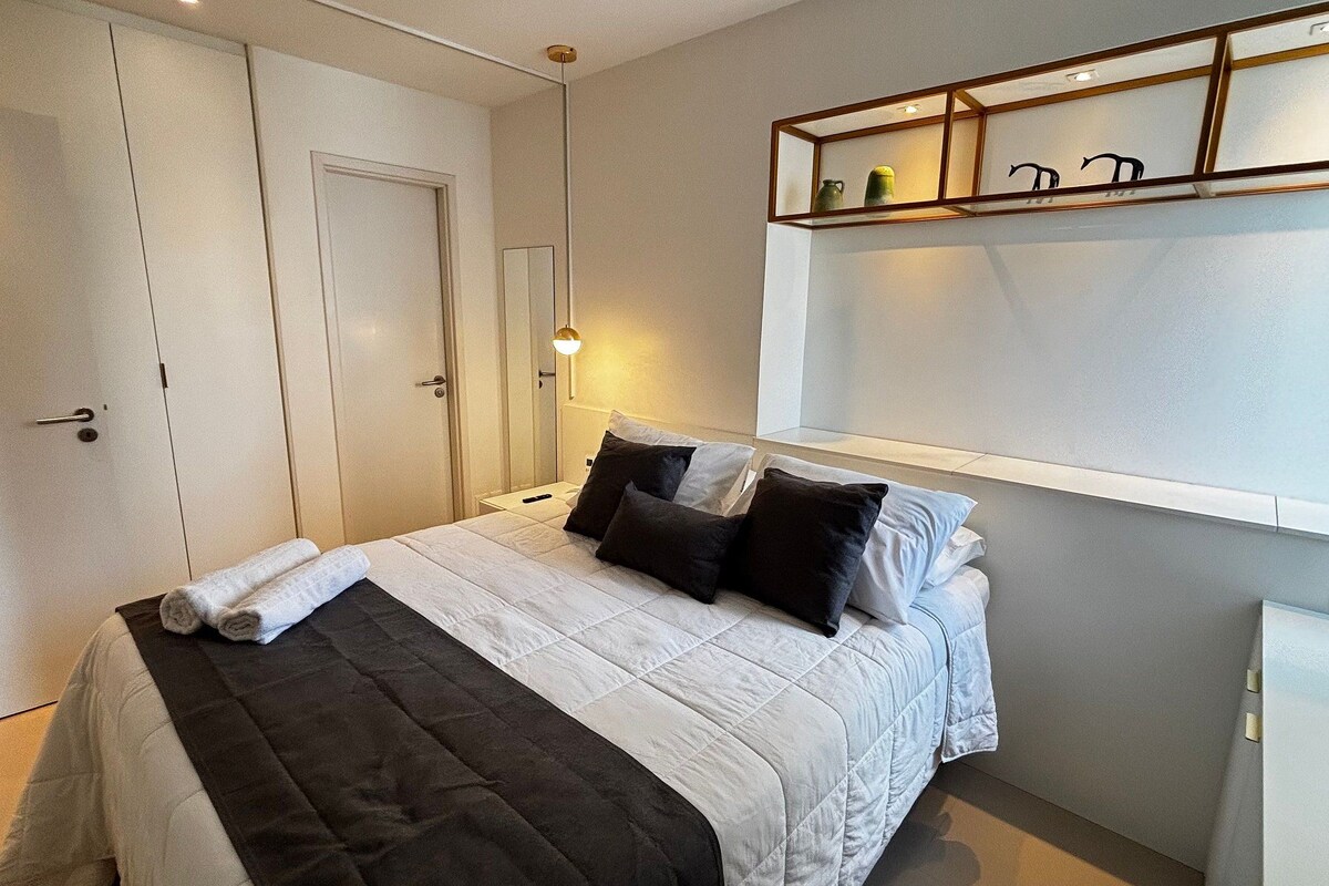 A neatly made bed with a white and black duvet, a bedside table with a lamp, and a shelf with decorative items.