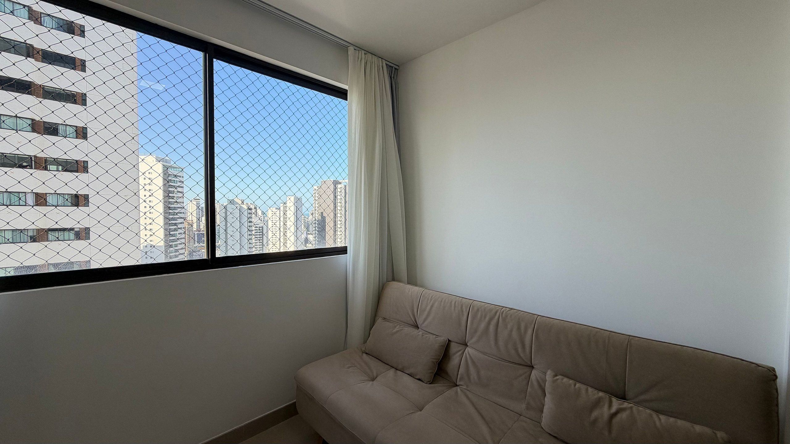 A room with a beige sofa, a window with a view of tall buildings, and a chain-link fence.