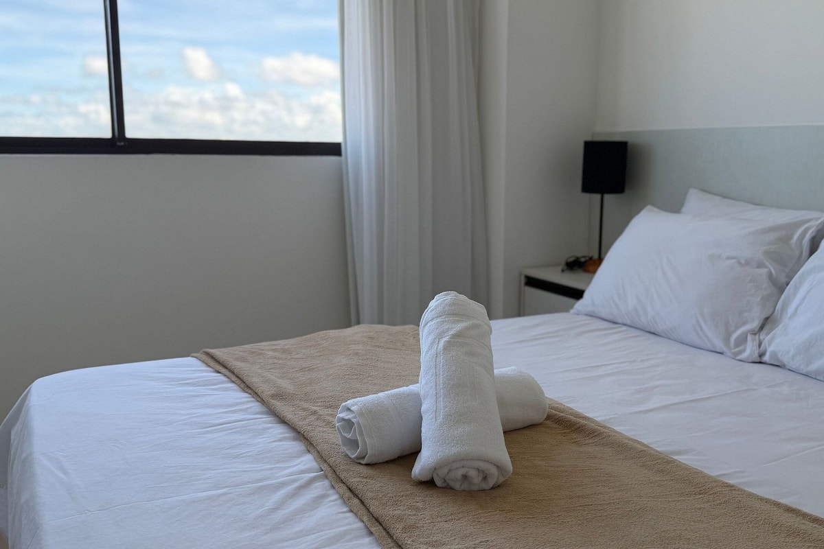 Habitación de hotel con cama, ropa de cama blanca, manta color beige y vista a la ciudad por la ventana.