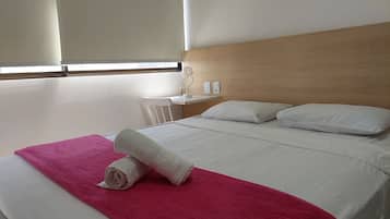 Una habitación de hotel con cama, cabecera de madera, mesita de noche y ventana con persianas.