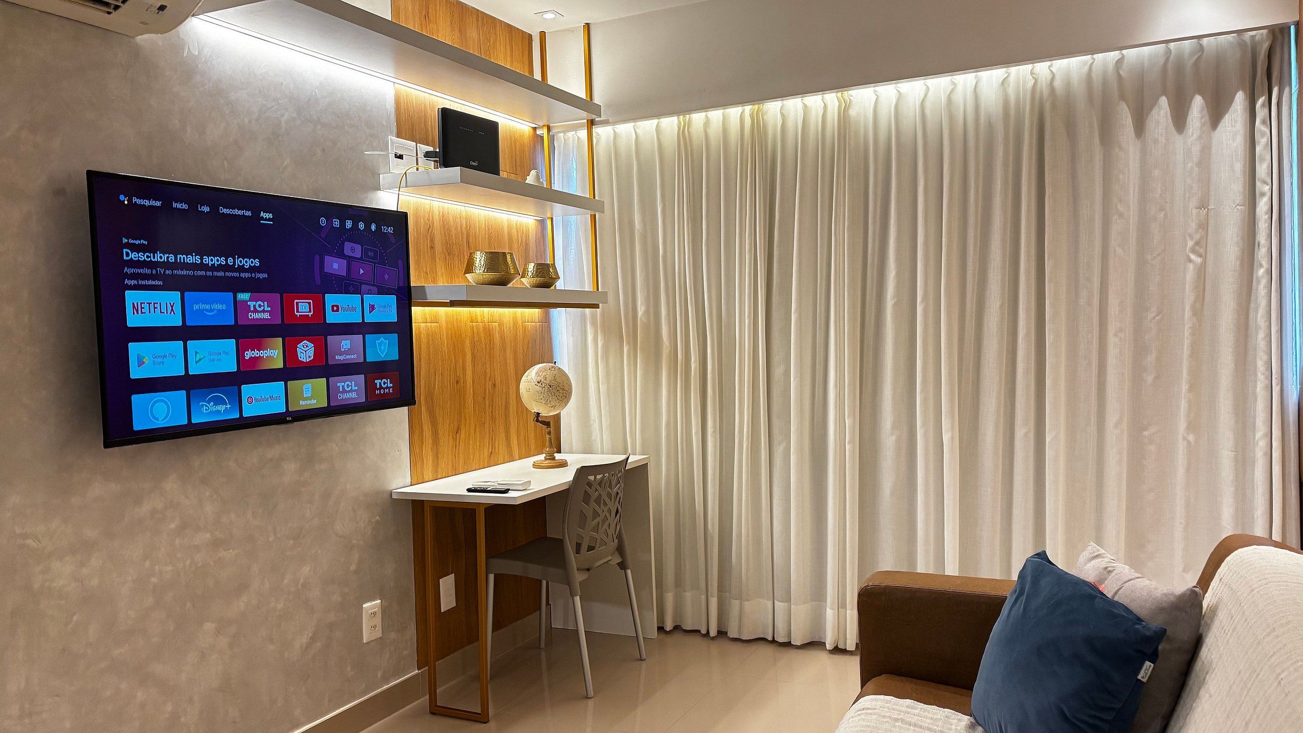 Una sala de estar moderna con un televisor montado que muestra varias aplicaciones, un escritorio con una silla y un amplio ventanal con cortinas.