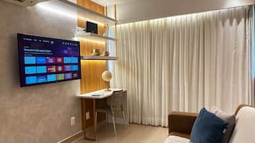 Een moderne woonkamer met een tv aan de muur waarop verschillende apps worden getoond, een bureau met een stoel en een groot raam met gordijnen.