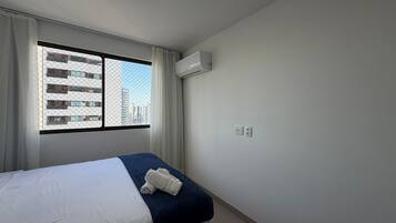 A hotel room with a bed, a window with a view of buildings, an air conditioning unit, and a power outlet.