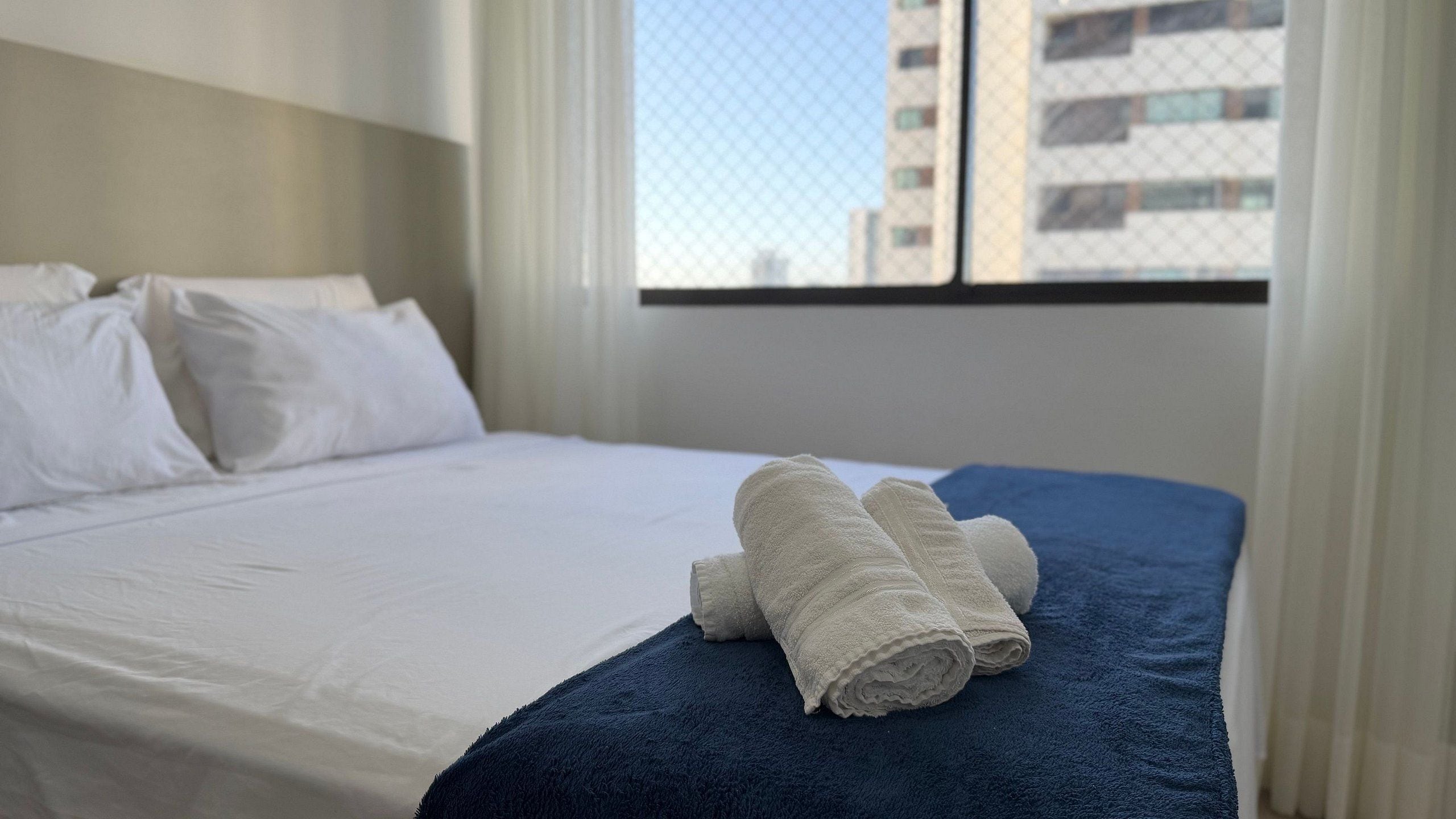 Een hotelkamer met een opgemaakt bed, een raam met uitzicht op gebouwen, en een blauwe deken op het bed.