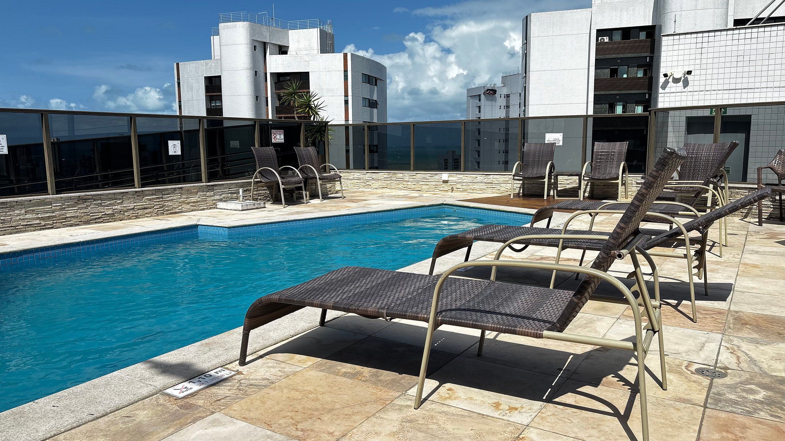 Una piscina en la azotea con sillones reclinables, un muro de piedra y edificios modernos al fondo.