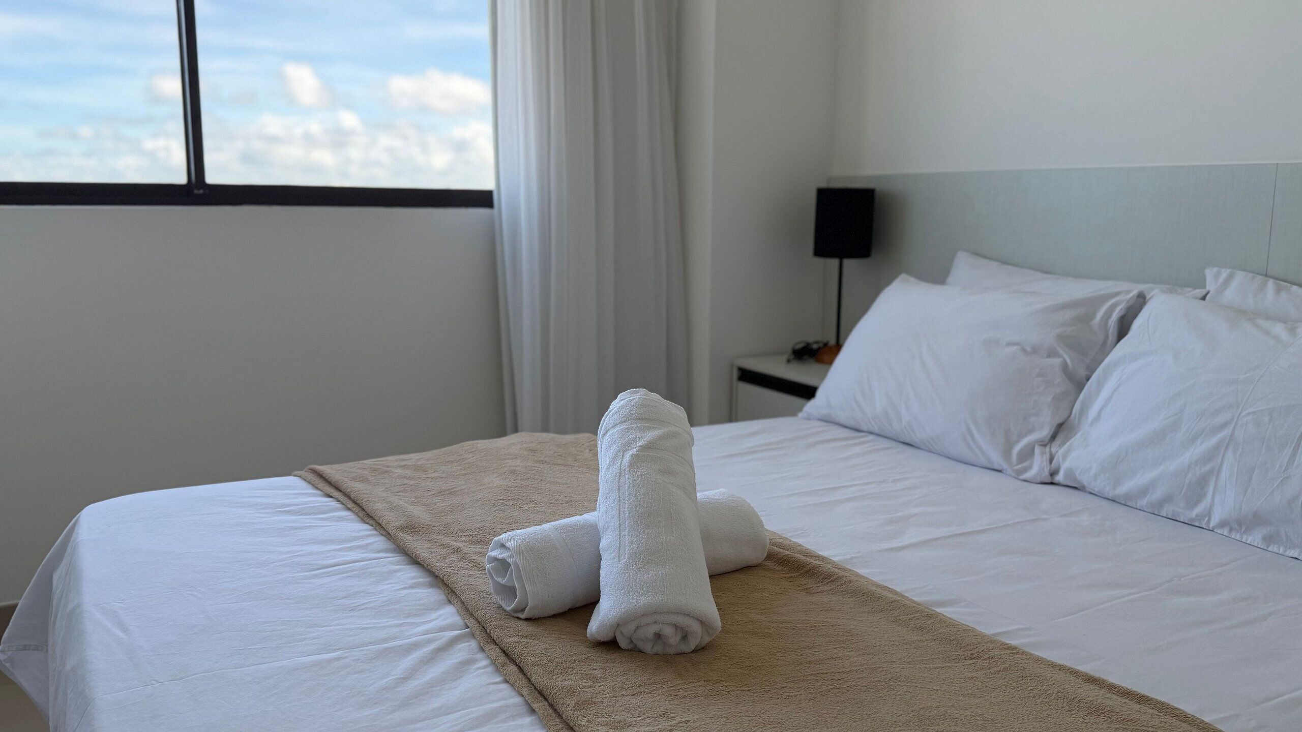 A hotel room with a bed, white linens, a beige throw blanket, and a view of the city through the window.