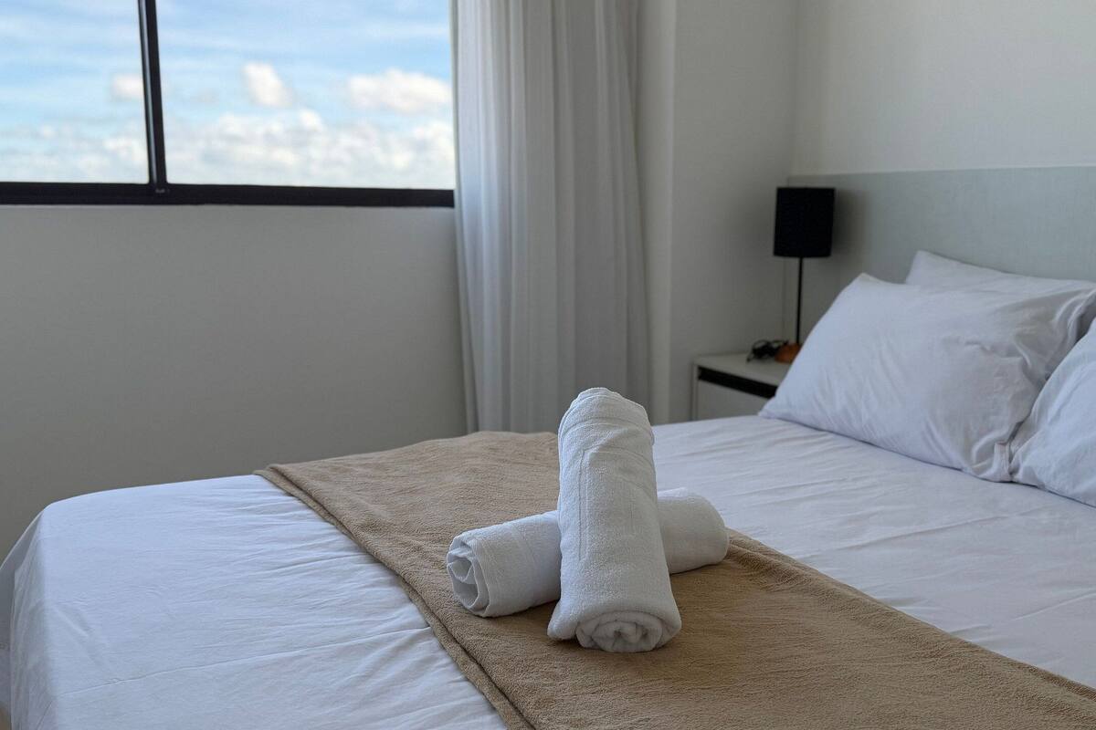 A hotel room with a bed, white linens, a beige throw blanket, and a view of the city through the window.