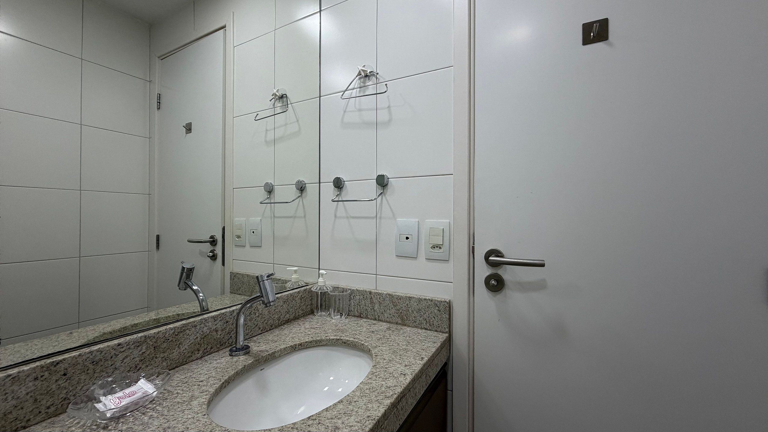 Um banheiro com bancada de granito, pia, espelho, porta-toalhas e porta com puxador.