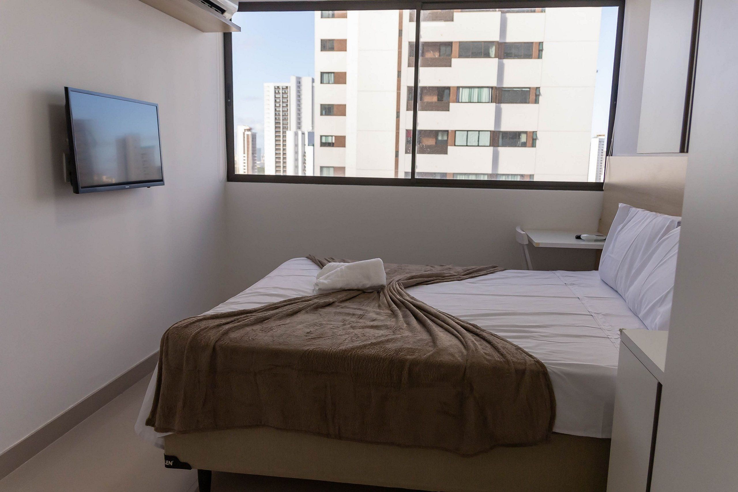 A hotel room with a bed, a bedside table, a TV mounted on the wall, and a window with a view of buildings.