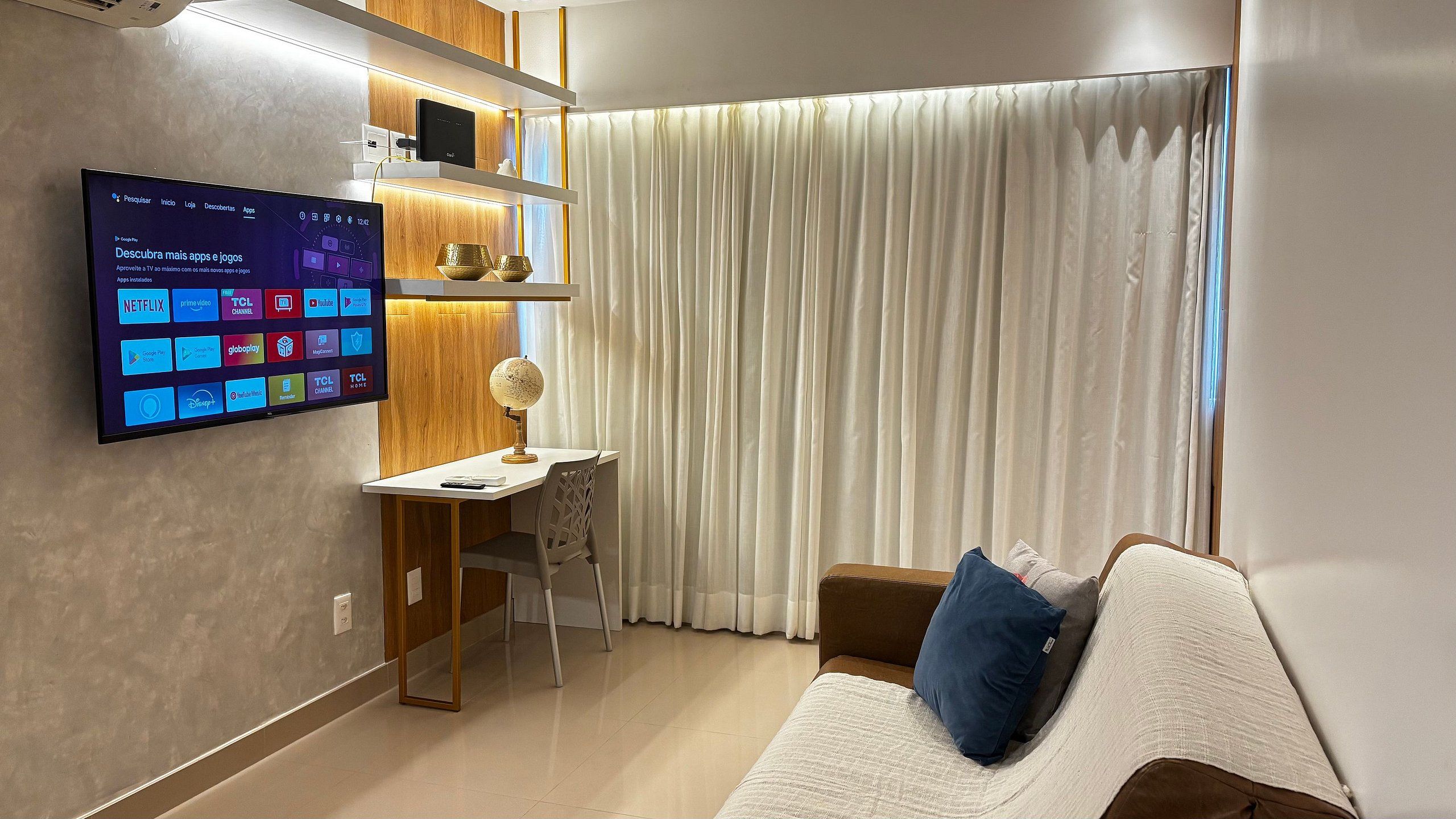 Una sala de estar moderna con un televisor de pantalla plana montado en la pared, un escritorio de madera con un globo terráqueo, un sofá marrón con un cojín azul y una ventana grande con cortinas blancas.