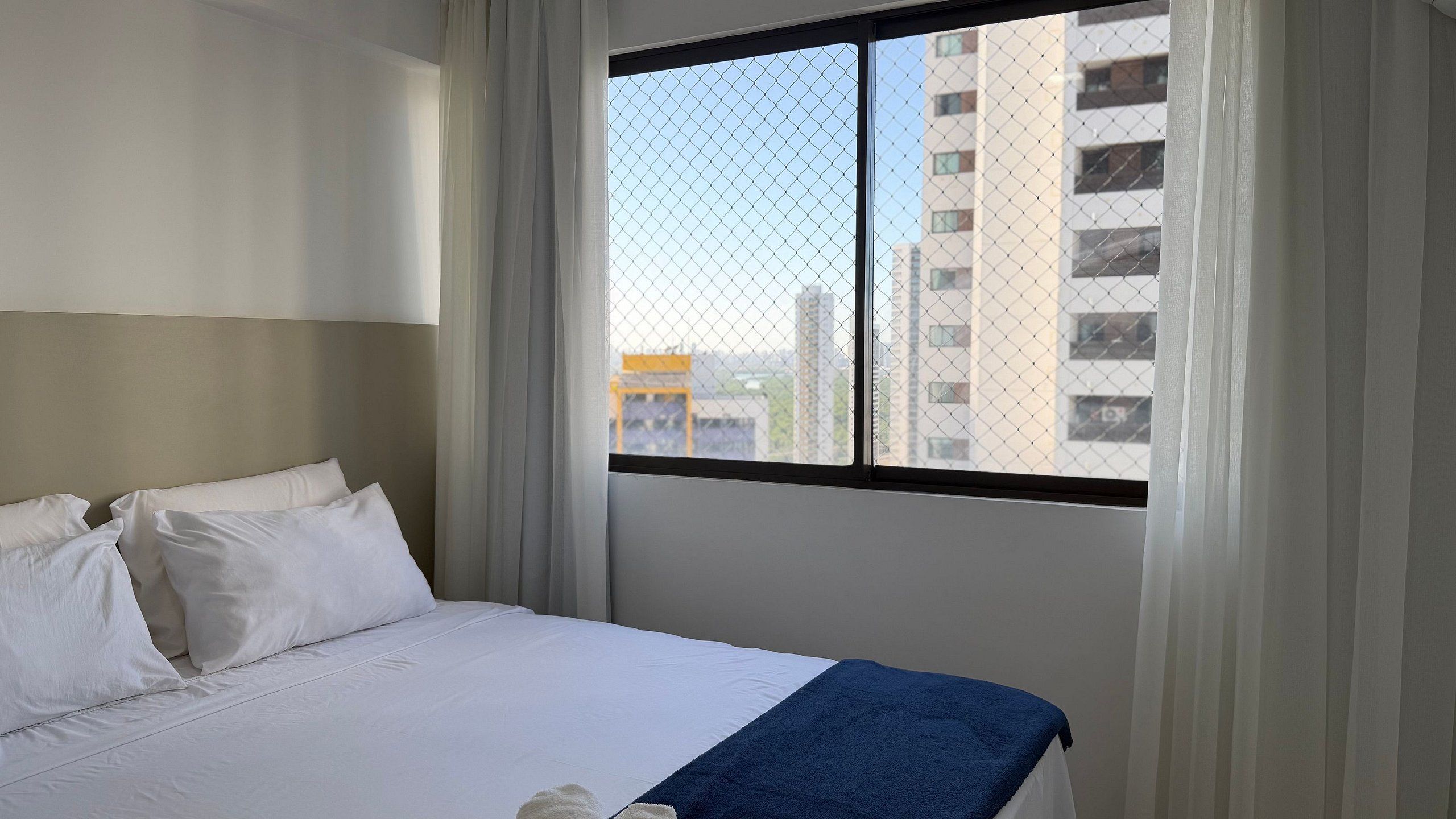 A hotel room with a bed, white curtains, a window with a view of buildings, and a blue towel on the bed.