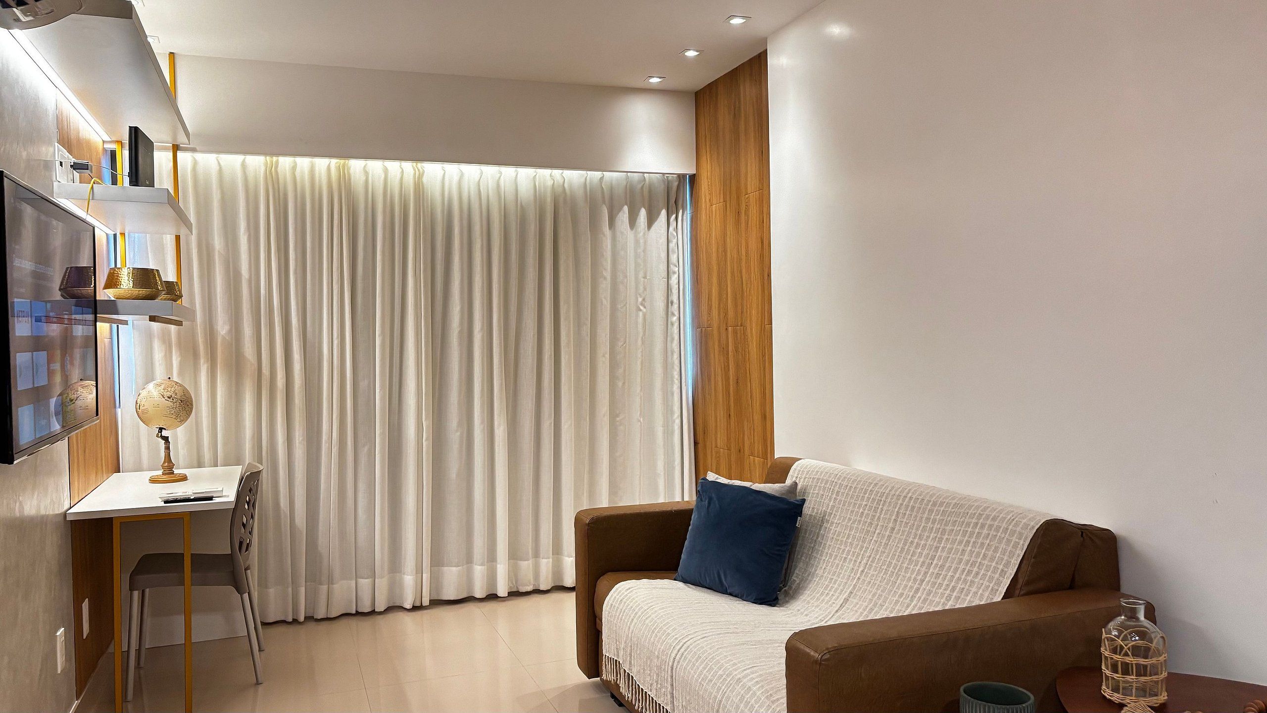 A modern living room with a sofa, a desk, a globe, and a large window with curtains.