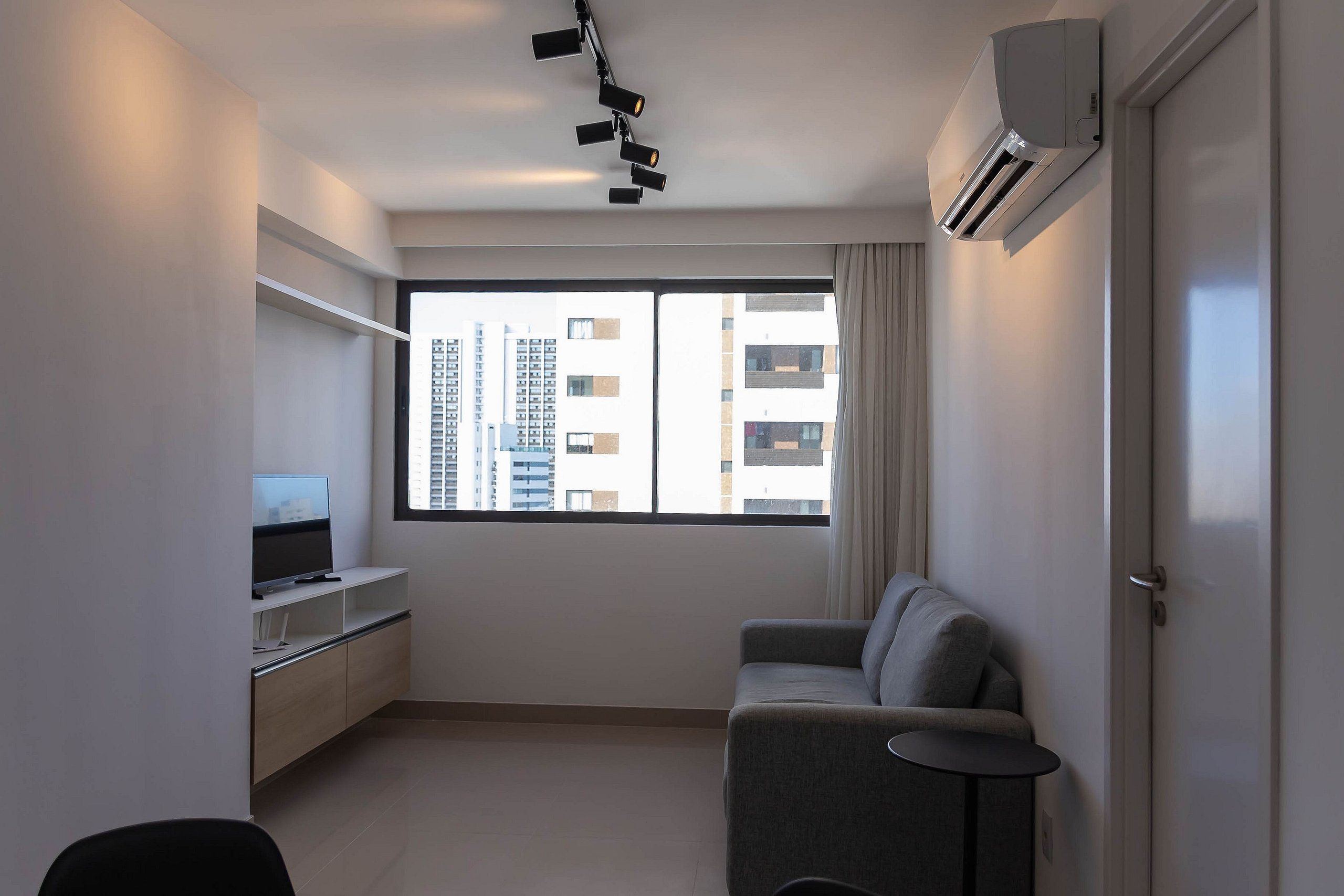 Un salon moderne avec un canapé, un téléviseur posé sur un meuble, un climatiseur fixé au plafond et une grande fenêtre donnant sur la ville.