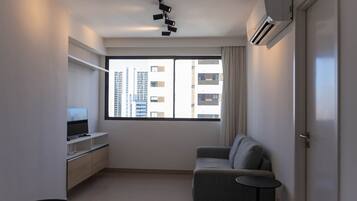 Un salon moderne avec un canapé, un téléviseur posé sur un meuble, un climatiseur fixé au plafond et une grande fenêtre donnant sur la ville.