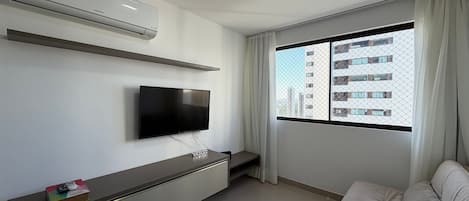 A hotel room with a flat-screen TV mounted on the wall, a sofa, a small table with a remote control, and a window with a view of buildings.