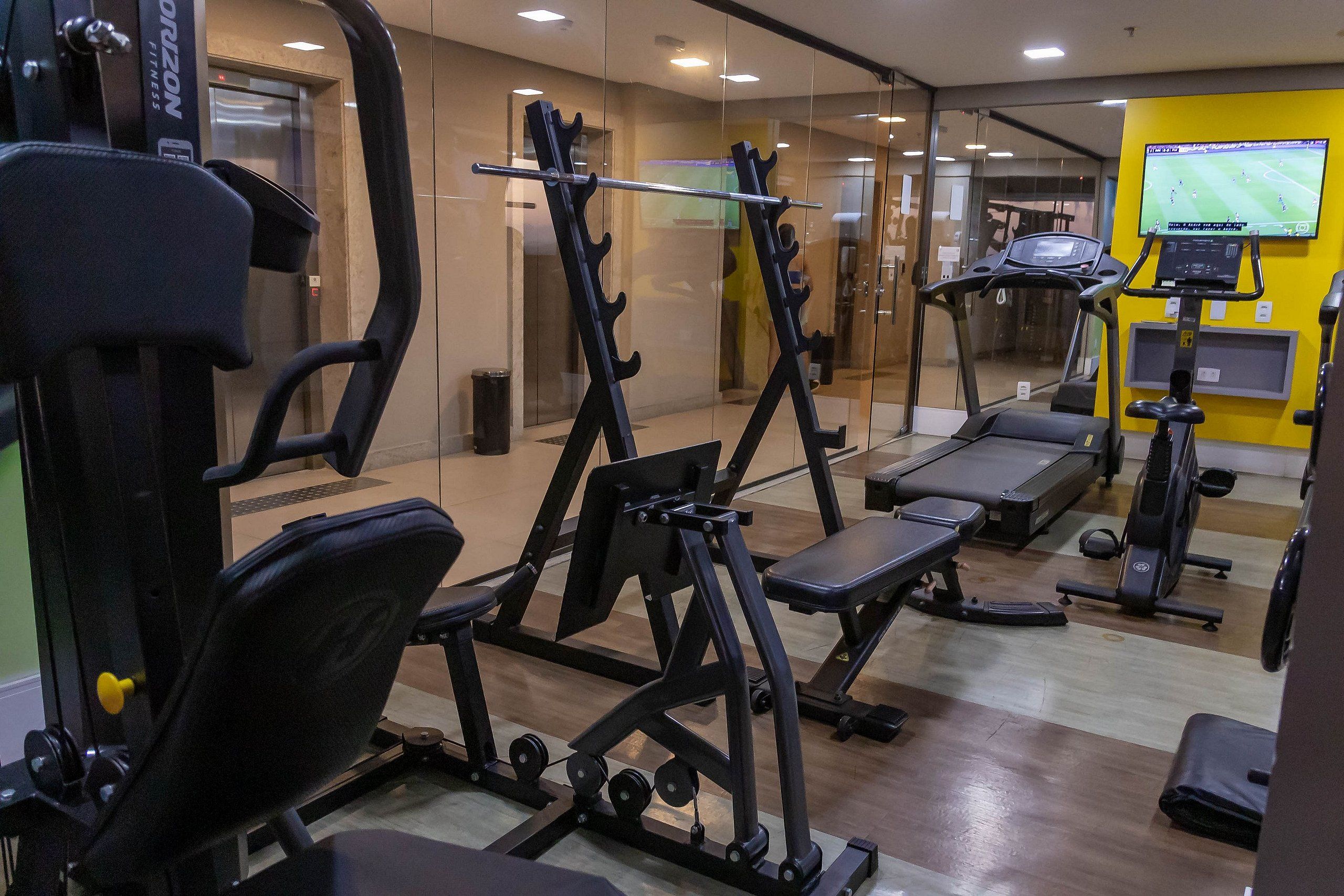 Un gimnasio con equipo de ejercicio, cinta de correr y una pantalla mostrando un evento deportivo.