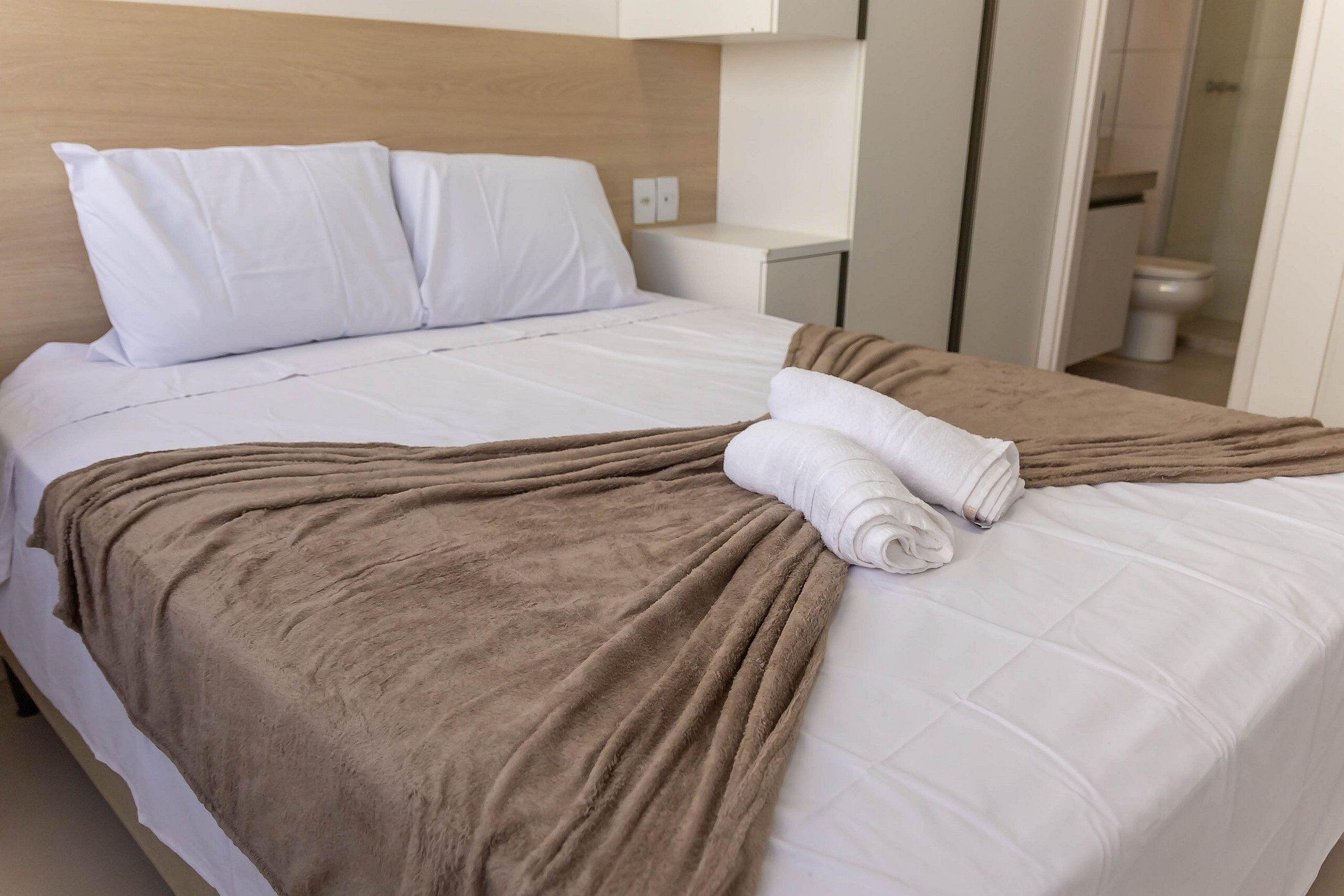 A neatly made bed with a brown blanket and white pillows, a white bedside table, and a view of a bathroom.