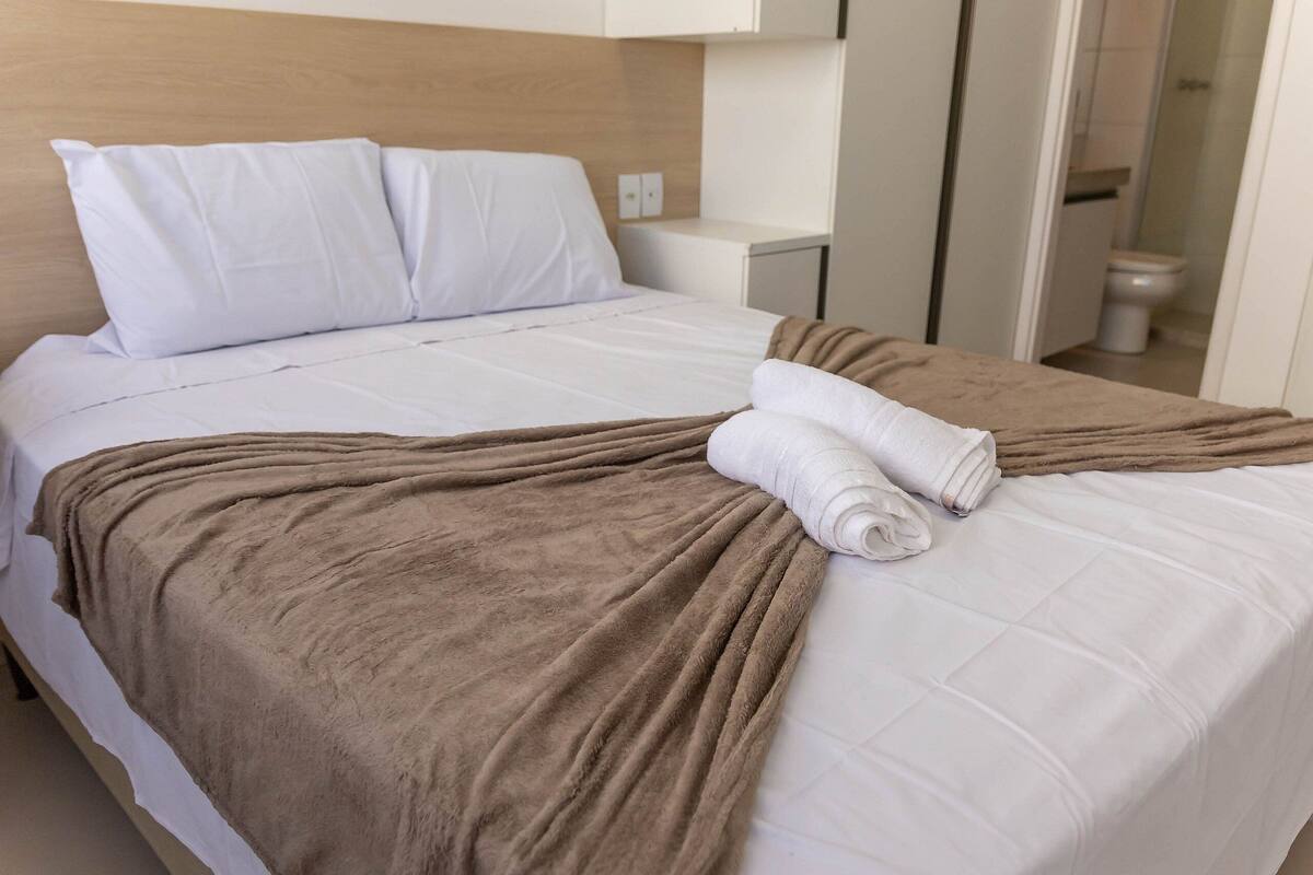 A neatly made bed with a brown blanket and white pillows, a white bedside table, and a view of a bathroom.