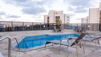 Una piscina en la azotea con sillones de descanso, vistas despejadas del paisaje urbano y un edificio moderno al fondo.