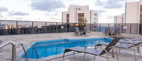 A rooftop pool with lounge chairs, a clear view of the cityscape, and a modern building in the background.