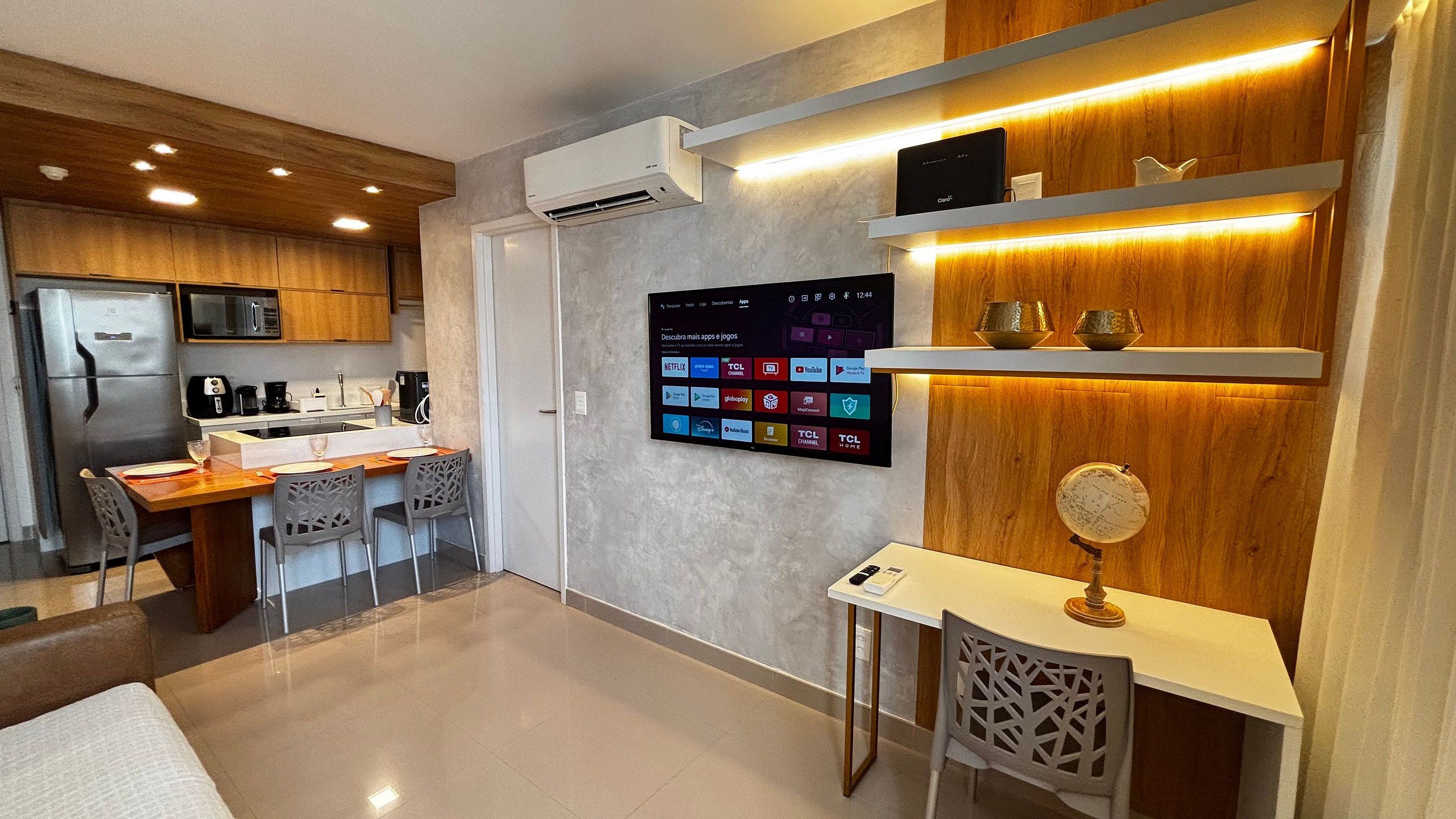 Una cocina moderna con aire acondicionado integrado, un comedor con mesa y sillas de madera, y un televisor fijado a la pared mostrando varias aplicaciones.