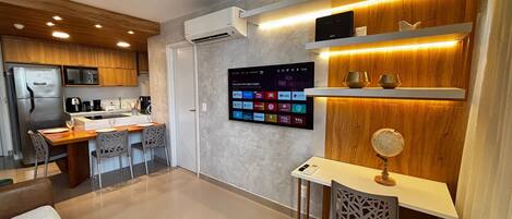 A modern kitchen with a built-in air conditioner, a dining area with a wooden table and chairs, and a wall-mounted TV displaying various apps.