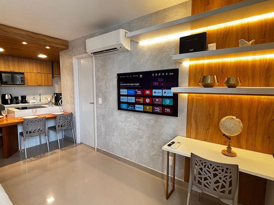 Uma cozinha moderna com ar condicionado integrado, uma área de refeições com mesa e cadeiras de madeira e uma TV na parede exibindo diversos aplicativos.