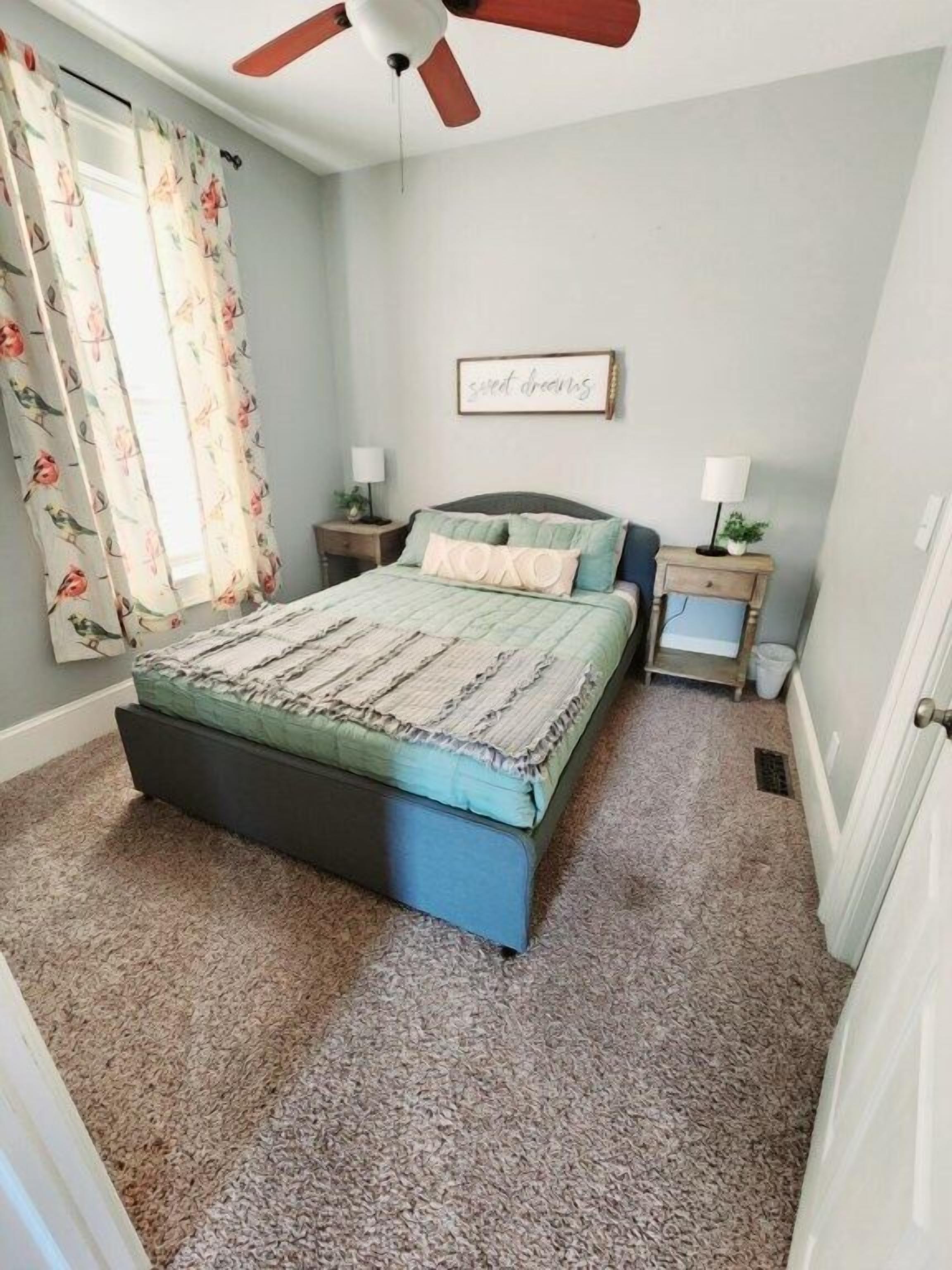 Quarto com cama, duas mesas de cabeceira, ventoinha no teto, janela com cortinas florais e placa fixada na parede.
