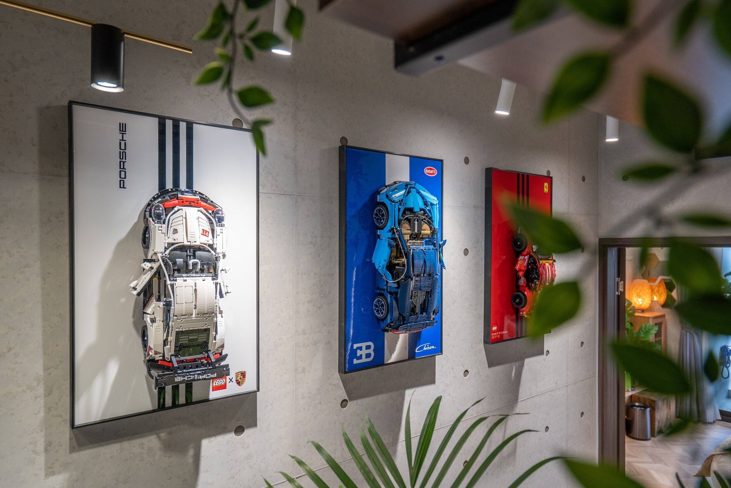 Drei Fahrzeugmodelle werden an einer Wand mit modernem Innenraum präsentiert, inklusive einer Pflanze und einer Tür zu einem anderen Raum.