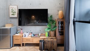 Ein modernes Wohnzimmer mit großem Flachbildfernseher, Couchtisch aus Holz, einer kleinen Pflanze und einer Küchenzeile inklusive Kühlschrank und Mikrowelle.