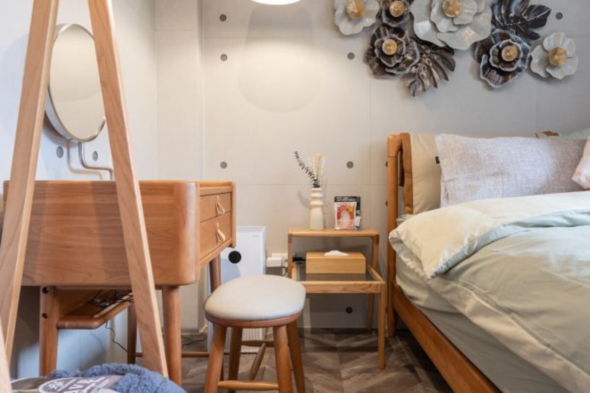A bedroom with a bed, a wooden desk, a stool, a mirror, and a wall-mounted flower decoration.