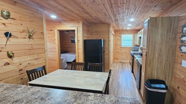 Interior de una cabaña de madera con una mesa de comedor, sillas, un refrigerador, un lavamanos y una ventana.