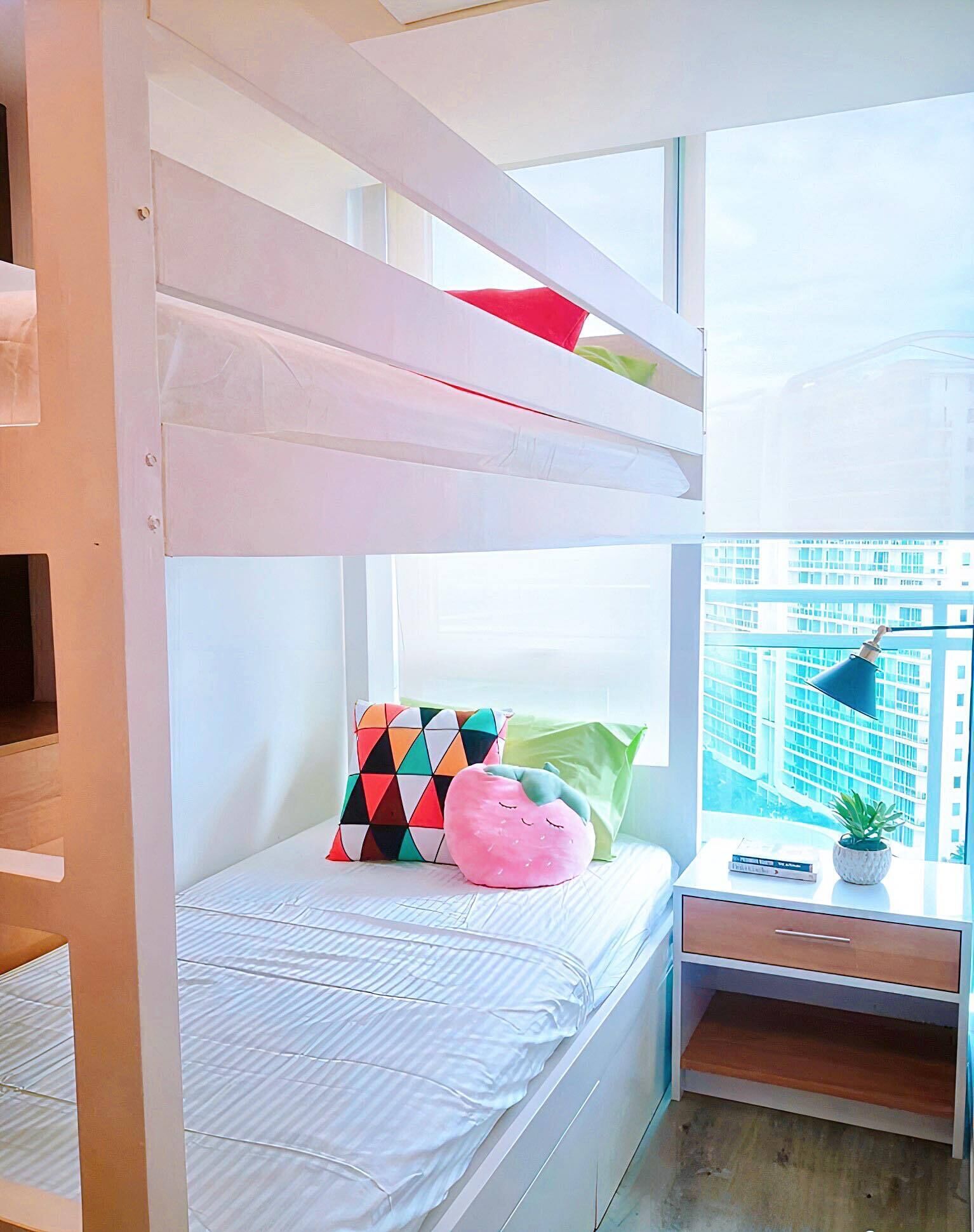 Un lit superposé avec une fenêtre, un bureau avec une lampe et une vue sur les immeubles.