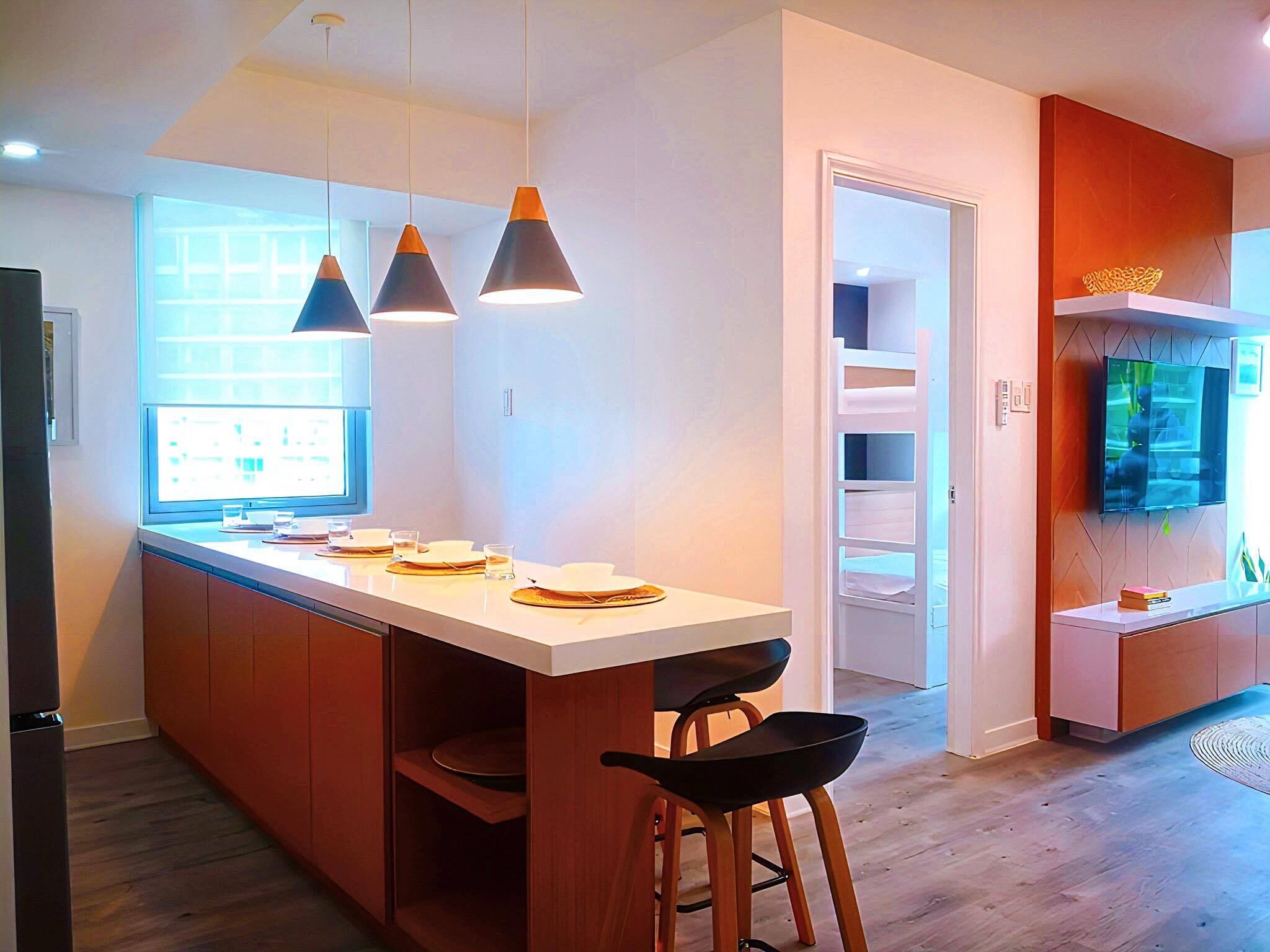 Una cocina moderna con iluminación colgante, un comedor con taburetes y un televisor empotrado.