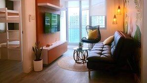 Un salon moderne avec un canapé, un téléviseur fixé au mur, une plante en pot et une vue sur la ville grâce à de grandes fenêtres.