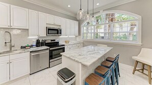 A modern kitchen with white cabinets, stainless steel appliances, a marble island, and a window with blinds. - Tuscan Tides - Custom Pool Home - powered by Island Real Estate (Holmes Beach)